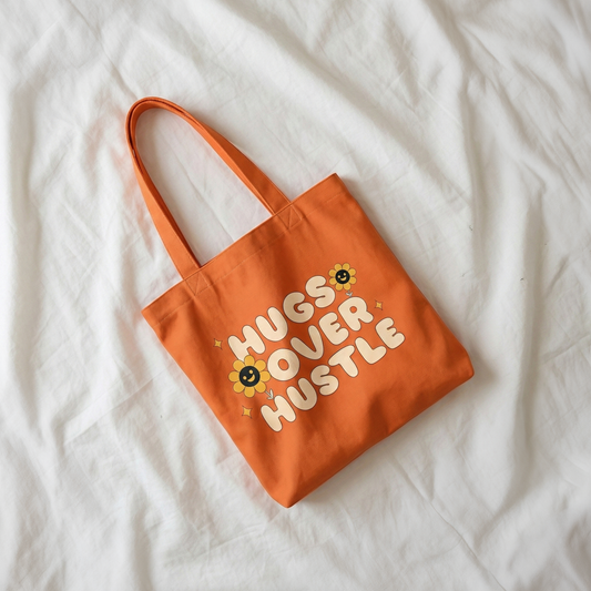 Orange canvas tote bag with “Hugs Over Hustle” quote placed on white bed sheet, cozy minimal lifestyle aesthetic