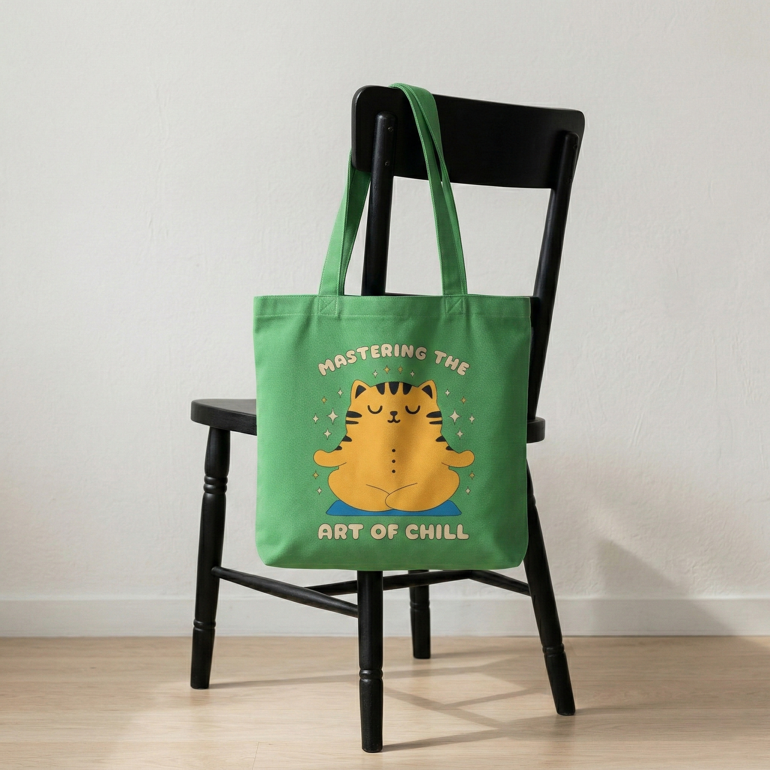 Green canvas tote bag with “Mastering the Art of Chill” meditating cat design placed on chair, cozy minimal home setting
